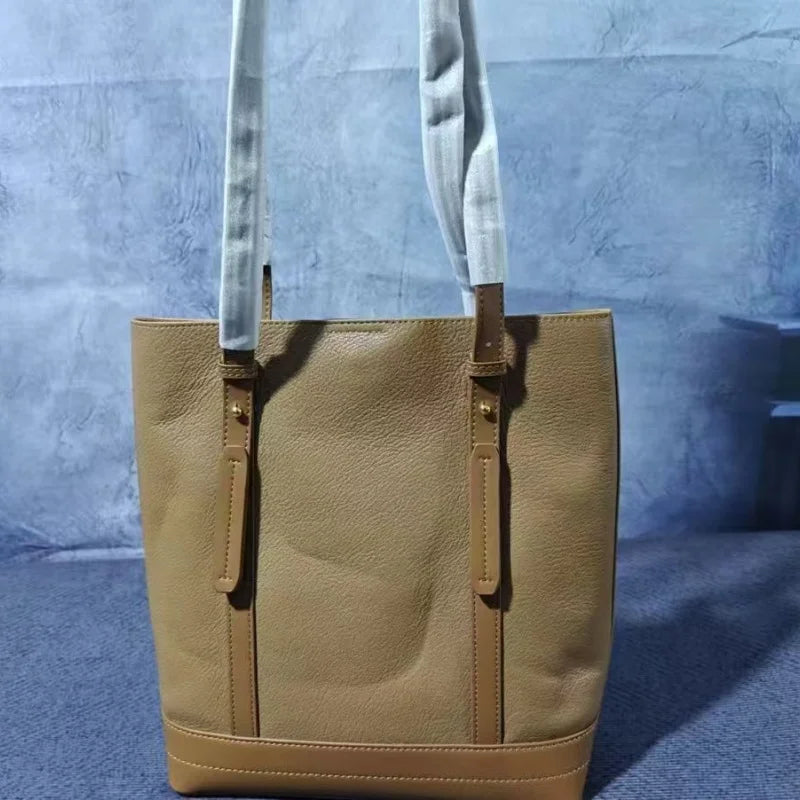 Leather vertical tote bag with riveted wide shoulder strap, decorative leather tag, stitched bottom panels, and large capacity.